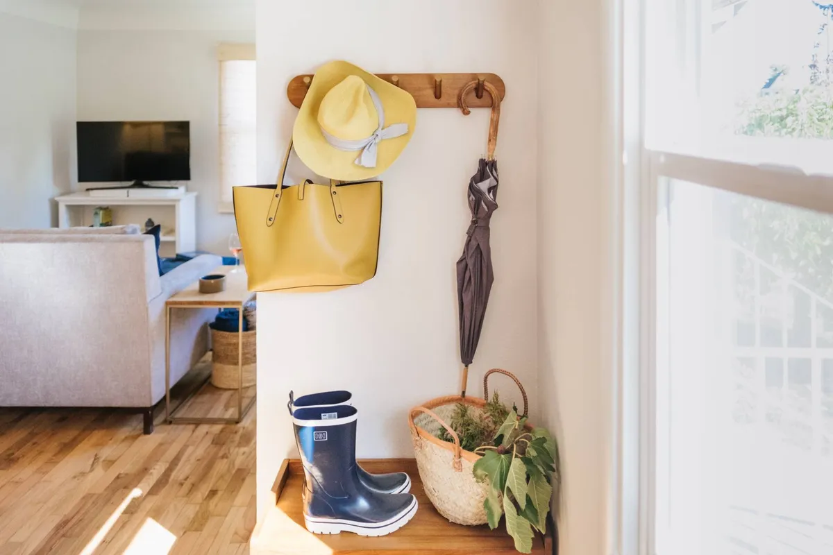 An entryway scene with a cheerful yellow handbag, a sun hat, a grey umbrella, blue rain boots, and a potted plant set beside a bench and hooks. The setup evokes a sense of arrival, readiness for outdoor adventures, and a practical place to store shoes and coats.