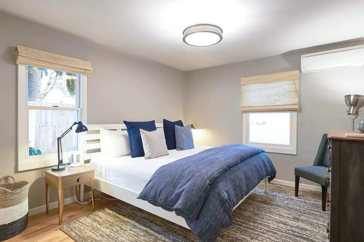 A king-size bed with blue and white bedding is centered in a serene, softly lit bedroom. Decorative pillows add a plush touch, while a nightstand with a lamp provides both style and function. There is a chair in the corner and a window letting in ample natural light, creating a tranquil and welcoming atmosphere ideal for rest.