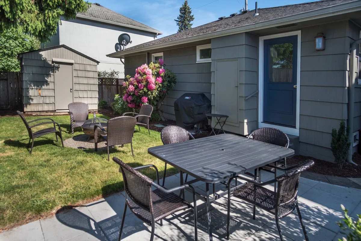 This image displays an inviting outdoor seating area set in a landscaped backyard, complete with a grill, dining table, and vibrant flowering bushes. The scene captures a relaxed atmosphere for social gatherings, outdoor meals, and enjoying the garden's natural beauty.