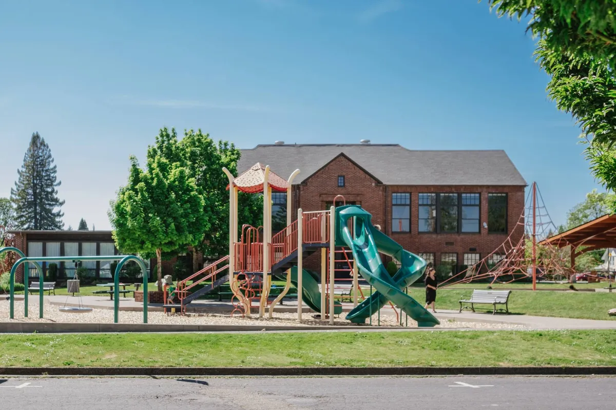 This exterior image captures a vibrant community playground with a green slide, surrounded by trees and benches, and a brick building in the background. Families can be seen enjoying outdoor activities such as picnicking, social gatherings, and playtime for children.
