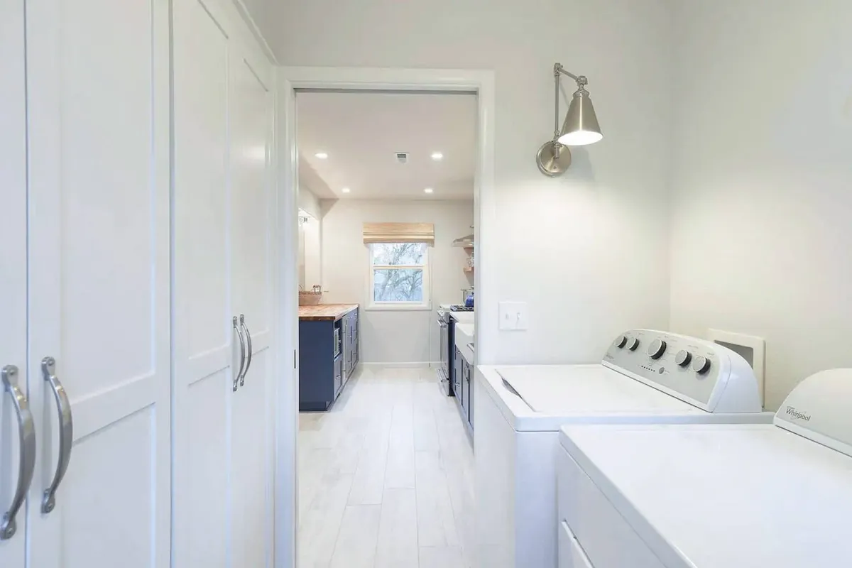 A practical, well-lit laundry area showing a full-size washing machine, with a doorway leading toward the kitchen. The scene emphasizes convenience and ease for longer stays.
