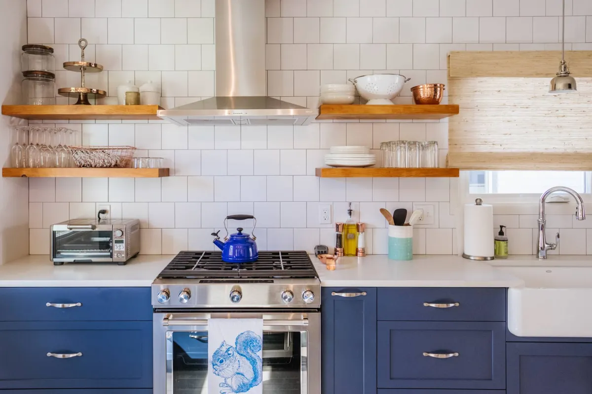 A warmly lit kitchen featuring a blue kettle on a stainless steel range, open shelving displaying kitchenware, and wooden accents throughout. The scene suggests active cooking and a welcoming space for gatherings.