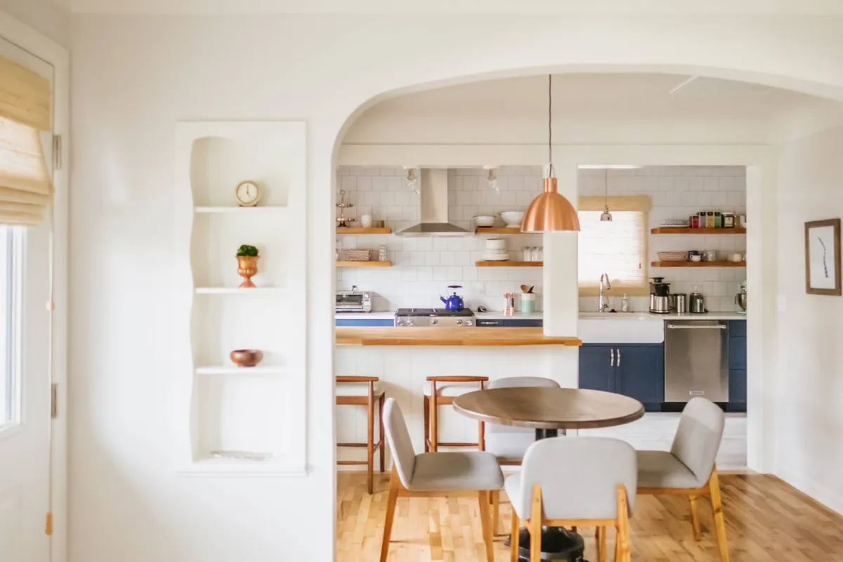 A cozy dining area is highlighted, featuring a wooden dining table beneath an eye-catching copper pendant light. The open kitchen layout in the background and shelves adorned with tasteful decor create a welcoming atmosphere suited for meals or casual gatherings.