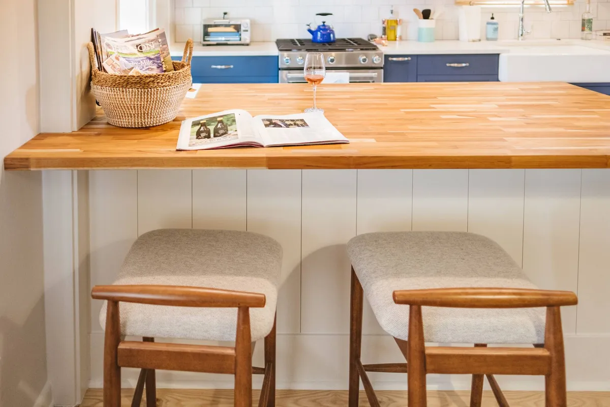 A close-up of a wooden eat-in bar counter with stylish bar stools, an open magazine, a wine glass, and blue cabinetry in the background. The scene radiates casual comfort and social interaction.