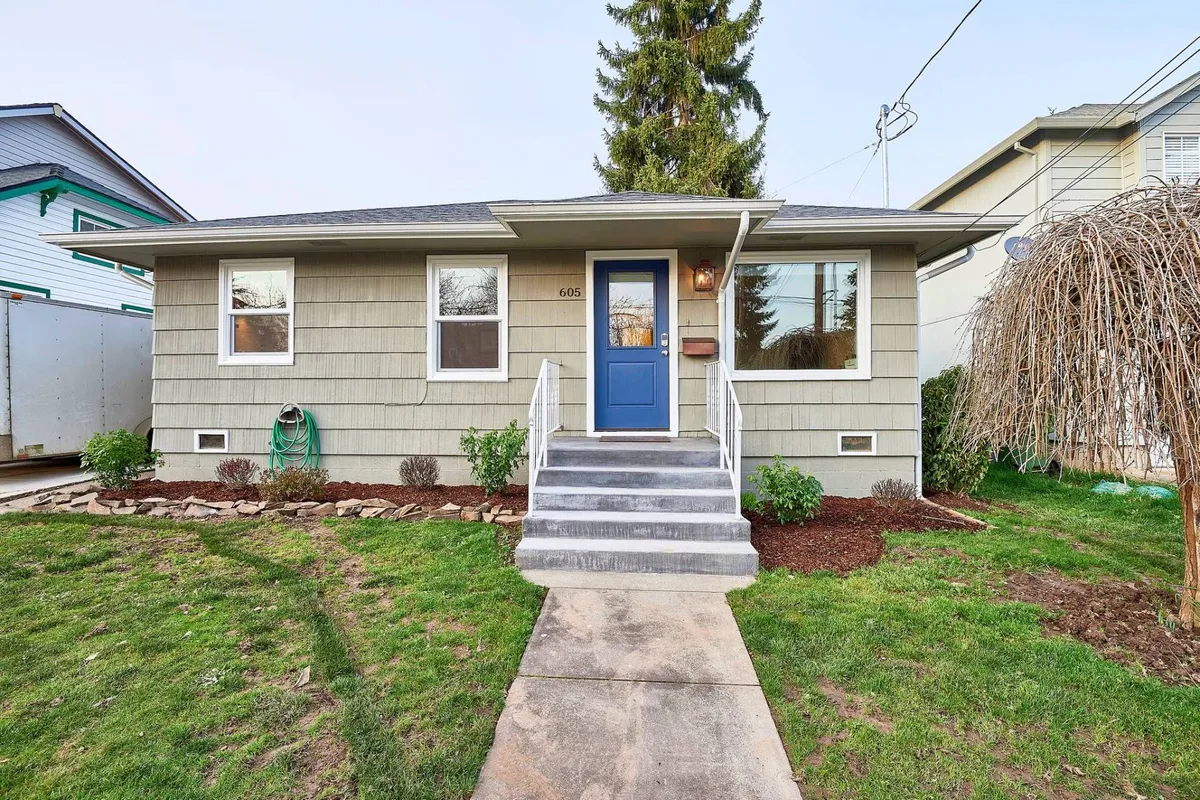 A welcoming blue front door framed by a lush, landscaped garden suggesting a charming, inviting cottage. Concrete steps lead up through greenery, and the inviting exterior hints at a warm retreat just moments from vibrant local attractions.