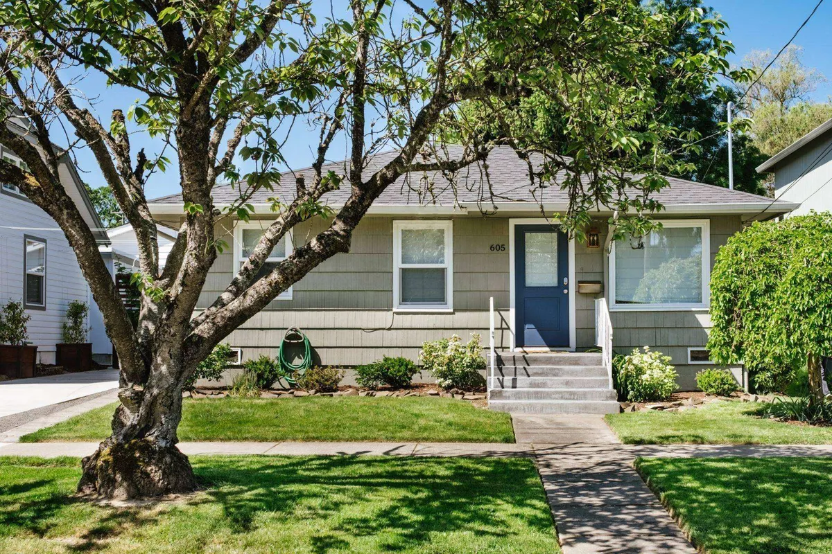 The image features the exterior of a charming single-family cottage with a welcoming front door, lush tree shading the steps, manicured garden area, and inviting path leading to the entrance. The front porch exudes serenity, and the garden hints at space for relaxing, outdoor meals, or watching the seasons change.