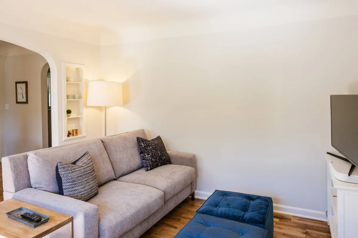 A welcoming living room is shown, featuring a plush sofa with velvet ottomans, a soft lamp casting a cozy glow, a built-in shelf with decorative accents, and a flat screen TV positioned for optimal viewing. The layout suggests a comfortable, inviting space ideal for relaxing or enjoying entertainment.