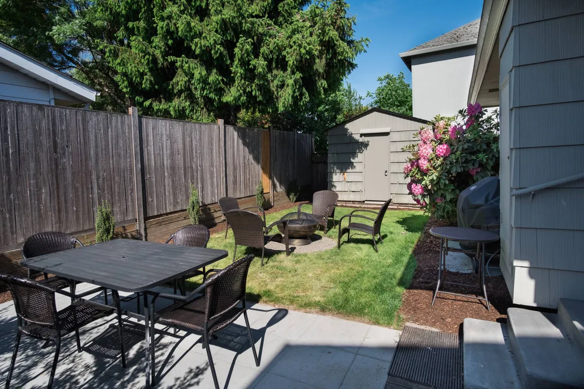 A warm, welcoming patio scene with a central table for outdoor dining, a fire pit, garden shed, and lush greenery. The image hints at sunset views and communal gatherings, encapsulating the spirit of shared alfresco experiences.