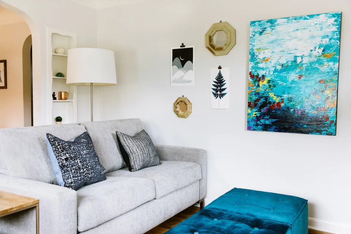 A cozy living area featuring a comfortable sofa, vibrant artwork above, an ottoman, and a soft lamp casting a warm glow. The scene is inviting and encourages relaxation, art appreciation, or thoughtful conversation, all while offering a peaceful, creative environment.